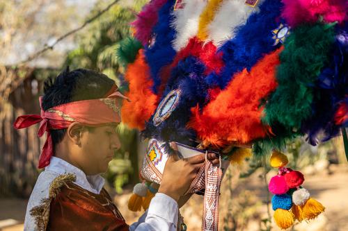 A man stands outside and holds and looks at a feather headdress, which is decorated with a variety of feathers. It is brightly colored, with blue, green, orange, pink, red, and white feathers. 