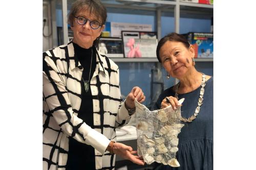 Two women smile, holding up a square piece of felt art, gauzy white with clumps of opaque white pieces and strands of brown fiber.