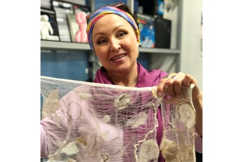 A woman in purple sweater and colorful headbands holds up a piece of fabric art.