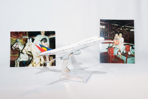 Display of two photographs and a model airplace, with a variation of the Filipino flag on the tail, on a white backdrop. The photos both depict a young boy and adult, on the left riding a wooden horse, and on the right standing on a mechanical airplane ri