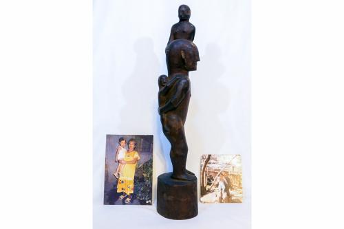 Display of two photos and a wooden sculpture of a person holding one child on its back and another standing on its shoulder. Old family photos show young boy with elders.