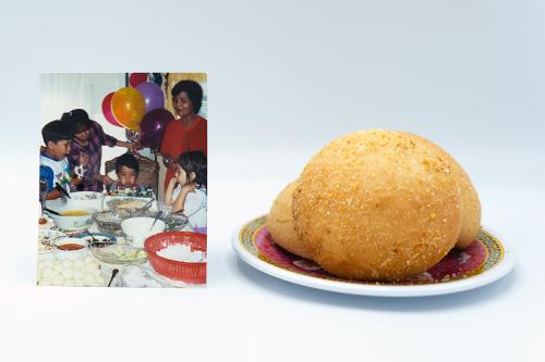 Family photo of a child’s birthday party, with a cake and many other dishes of food on the table. Next to the photo, a small plate containing three bread rolls.
