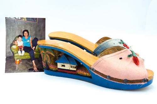 On the left, photo of a child and adult sitting together in a green armchair. On the right, a pair of wooden heeled sandals. The sides of the heels are carved with the scene of a white house with blue roof and a palm tree.