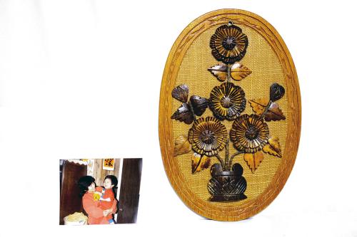 A photo of a mom holding up a todder, both dressed in red. To the right of the photo, a brown wooden and woven artwork depicting a vase of flowers.