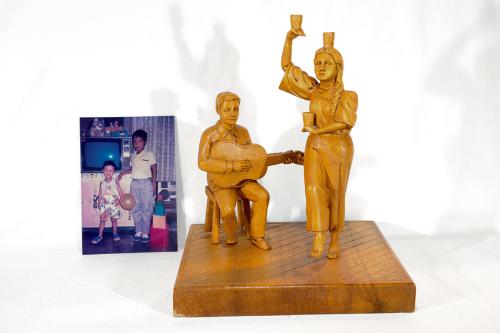 A photo of two young boys standing in front of a TV. In front of the photo, a wooden sculpture of a man, seated, playing guitar while a woman danced holding cups balanced on her head and hands.