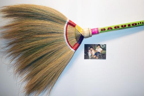 A handmade grass broom, woven where the strands are gathered with a red, yellow, and black design. The green handle reads: BAGUIO. A photo sits next to it of two children.