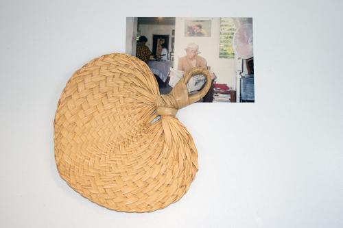 A tan woven fan with a loop handle. Under it, a photo of an elder man sitting and reading the newspaper inside a house.