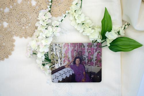 A photo of a woman in purple dress leanings on the back of a couch, draped with white lace. Underneath the photo, an arrangement of white plastic jasmine flowers, green leaves, and tan doilie lace. 