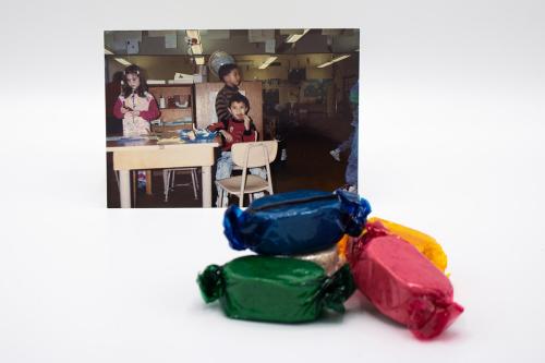 A small stack of candies wrapped in blue, green, pink, and orange wrappers, in front of a photo of kids in a classroom, one eating a snack.