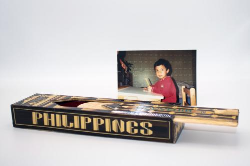 Wooden rectangular container with a top that slides open. The black and nature design reads PHILIPPINES on the side. Behind it, a photo of a boy at a desk, holding a pencil.