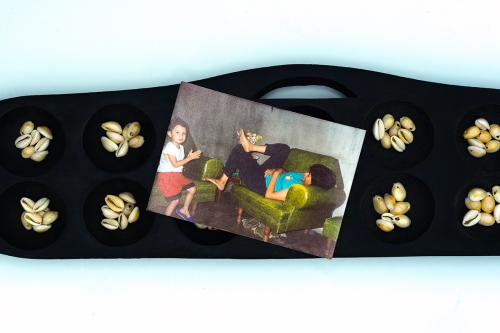 A photo of a child and adult on green armchairs rests on top of a black wooden game board, with cups and a handle carved into each. Each cup holds seven small cream-colored shells.