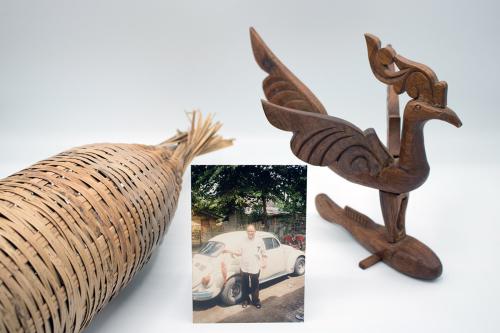 In the center, photo of an elder man in front of a white VW Beetle. To the left, a tan woven reed fish trap. To the right, a darker brown wooden carving of a bird.