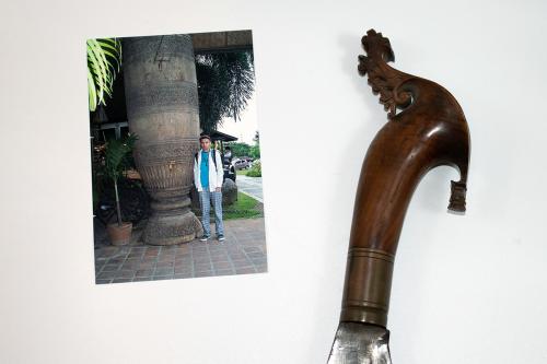 In the foreground, a stereoscope card with doubled image of a group of people in a courtyard. The photo rests against a tan wooden instrument. In the background, photo of a young man holding the same type of instrument, with a rectangular body, held like 