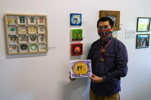 A man in embroidered face mask and gold pendants on necklaces holds up a small abstract painting in yellow and purple. Behind him on a gallery wall, other paintings and display cases.