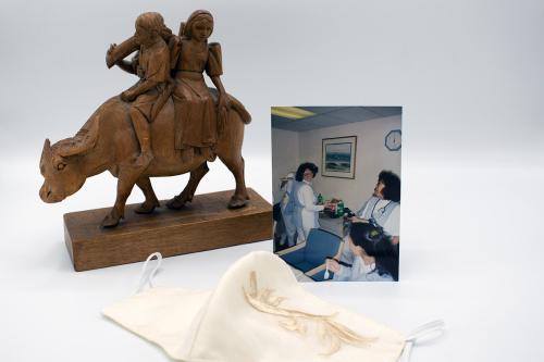 Display of a brown wooden carving of two people riding an ox, a white embroidered face mask, and a photo of women in nurse uniforms, all on a white backdrop.