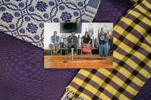 In the center, photo of four people seated on a stage, one speaking into a microphone. Under the photo, arrangement of three types of woven cloth: one purple and black, one purple and yellow plaid, and one white and blue with geometric pattern.
