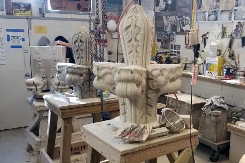 In a workshop cluttered with tools and work benches, we see three pieces of carved stonework each on its own table. The center one (the original) is dark, caked with dust. The one in front has almost the exact same design but is bright white and clean.