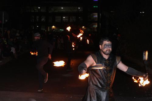 People dressed in all black parade down a dark street carrying or other flaming objects.