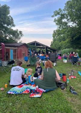 Adults and children sit on blankets and camp chairs on a green farm.