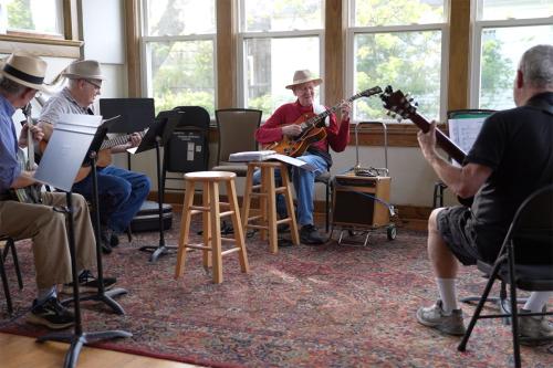 Four men sit in a circle in a practice room, each playing guitar. Next to the man in the center, Frank Hamilton, is a guitar amp strapped to a small dolly.
