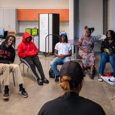 In a classroom, one person faces away toward a semicircle of teens, seated and laughing.