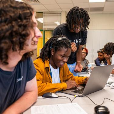 Teens crowd around laptops in a classroom, smiling.
