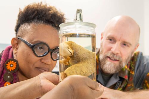 A Black woman and a white map both look closely at the lizard in a jar.