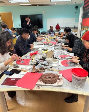 Around an indoor table, seated people work on clay faces on red paper.