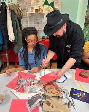 An man in black clothes and fedora painted on a sheet of red paper while a seated person watches.