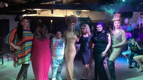Seven people in drag pose arm in arm instead a club, with colored lights and synthetic fog in the background.