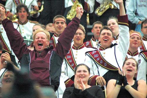 A man shouts and raises his arms in victory, with members of the marching band in uniform behind him cheering.