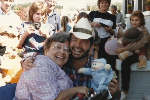 An older woman and a man, both sitting or kneeling, embrace and smile at the camera, with a crowd of people behind them. The man is holding up a Cabbage Patch Kid.
