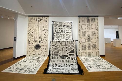 Art display in a gallery: Three long sheets of white paper, extending from the ceiling to spread across the floor, with geometric patterns in watery black ink.
