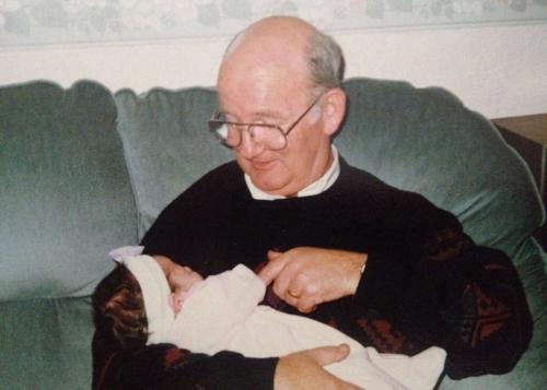 A man, seated on a couch, cradles a baby in a pink onesie. Old color photo.
