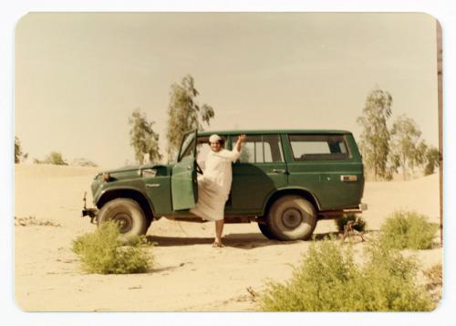 A person in white robe steps out of a green vehicle in a desert landscape.