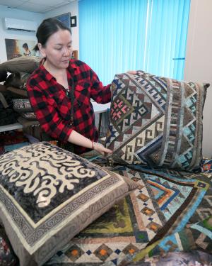 A woman holds up a square pillow, covered in an intricate multicolored embroidery. Below it is a stack of other embroidered pillows and textiles.
