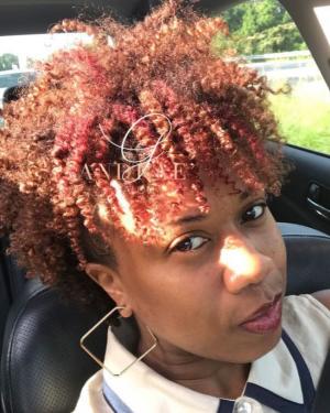 Person posing in the driver seat of a car with reddish, curly natural hair.