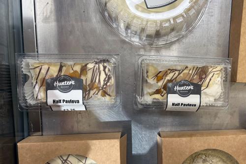 From above, two clear plastic containers labeled Half Pavlova. The creamy dessert inside is drizzled with chocolate. Other boxed desserts are half out of frame.