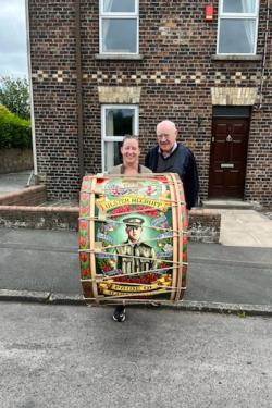 A young woman and elder man pose on a sidewalk in front of a brick building. The woman holds an enormous drums, painted with the portrait of a soldier on the side.