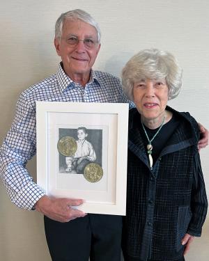 An elder man and woman pose arm in arm, holding artwork in a white frame. The illustration is the same at the beginning of this article: a young boy with two gold coins.