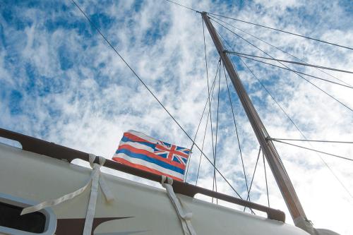 Looking up toward the mast of a boat and wispy white clouds in a blue sky, the Hawaiian flag flies: the UK Union Jack in a top corner and eight horizontal stripes in white, red, and blue.
