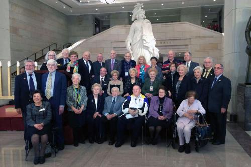 Roughly thirty elderly men and women in formal wear pose in three tiers in front of a Greek-style marble sculpture.