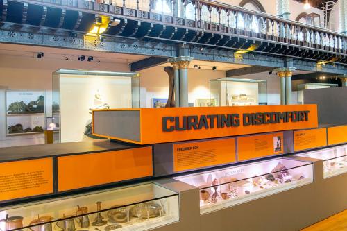 Two-story museum gallery with display cases and signage in bright orange, including the exhibition title: Curating Discomfort.