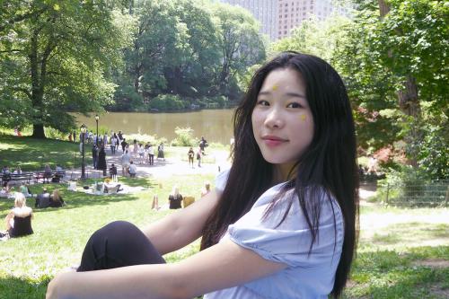 An Asian woman with long dark hair smiles, sitting on the ground in a park with a pond and buildings in the distance.