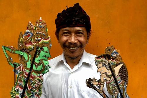 A man smiles at the camera, holding two intricately designed shadow puppets of human figures, controlled  by wooden sticks that run the length of their bodies. He wears a bright white dress shirt and traditional darkcolored peci headwrap. 