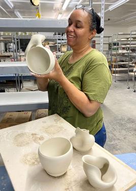 A woman holds up a ceramic bowl in progress. Other bowls, including one that appears crumpled, sit on her worktable. 