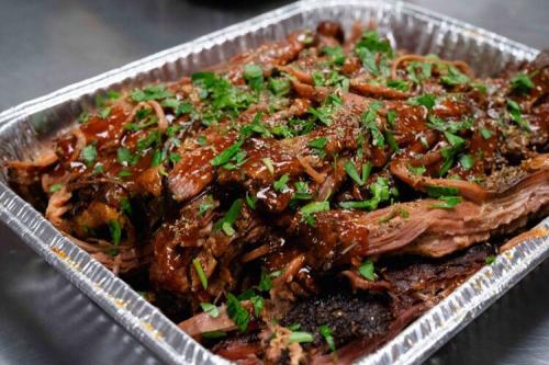 An aluminum catering dish filled with deep brown, glazed pulled chicken, topped with sliced parsley.
