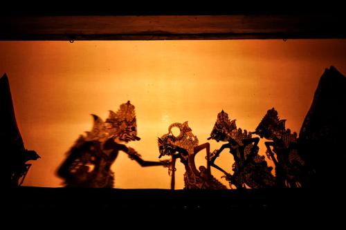 Silhouettes of four stylized human figures in shadow puppet form again a screen. On either side of them are space-shaped stationary scenery pieces.