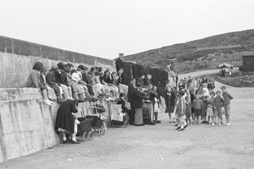 Children stand near and sit on what appears to be a cement seawall, surrounding a group of musicians performing. Of the instruments, we can only see a large bass drum and two fiddles. Black-and-white photo.