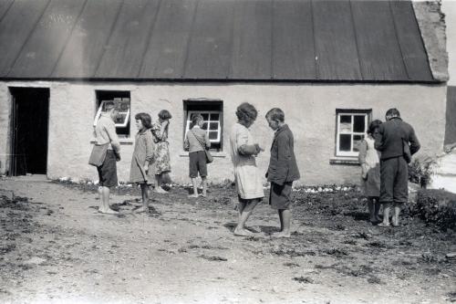 School kids stand in pairs, face to face, outside an old house. Black-and-white photo.